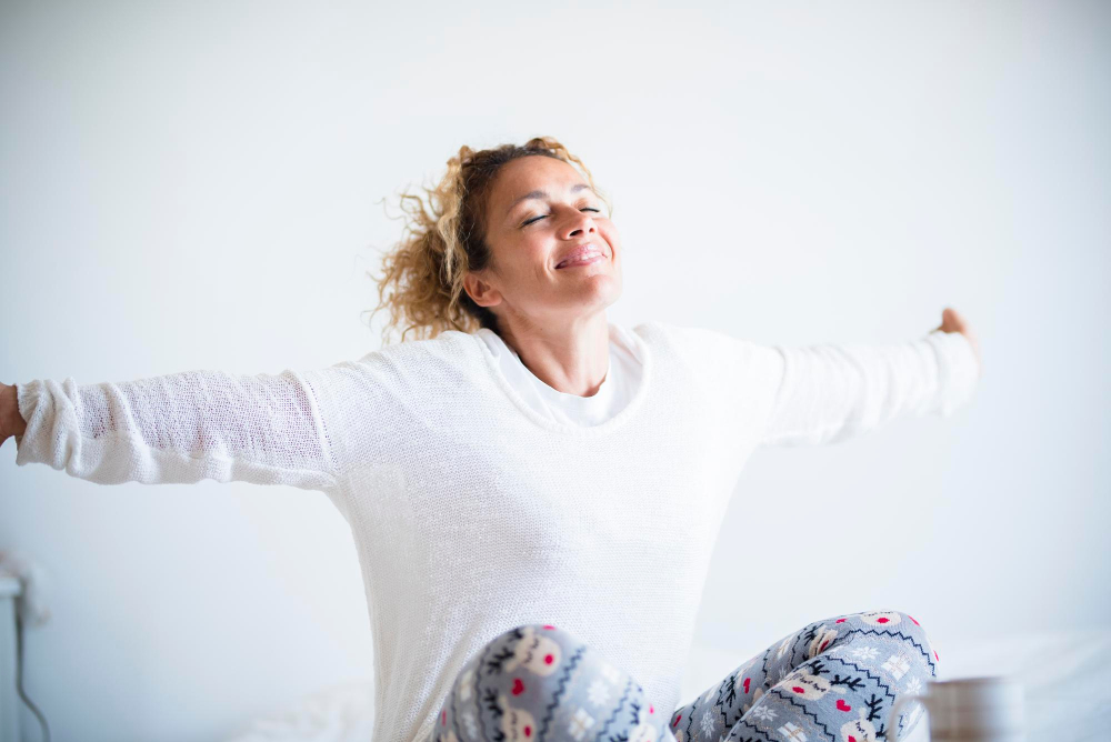 Eine Frau mit lockigen Haaren sitzt in ihrem Bett und streckt sich mit einem Lächeln und geschlossenen Augen wohlig aus, was einen entspannten und positiven Start in den Tag symbolisiert.