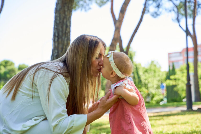 Eine Frau kniet in einem sonnigen Park und gibt einem Baby in einem rosa Kleid mit Stirnband einen liebevollen Kuss – ein bewegender Moment, der die Vorfreude und Bedeutung zeigt, Tante zu werden.
