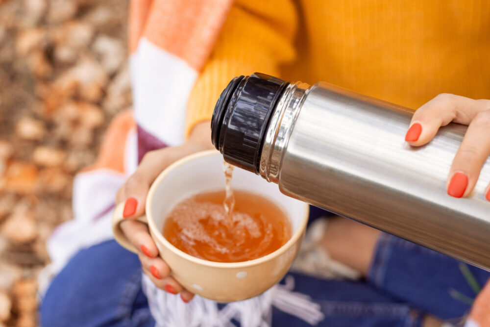 Aus einer silbernen Isolierkanne mit schwarzem Verschluss wird bernsteinfarbener Tee in eine helle Tasse gegossen.