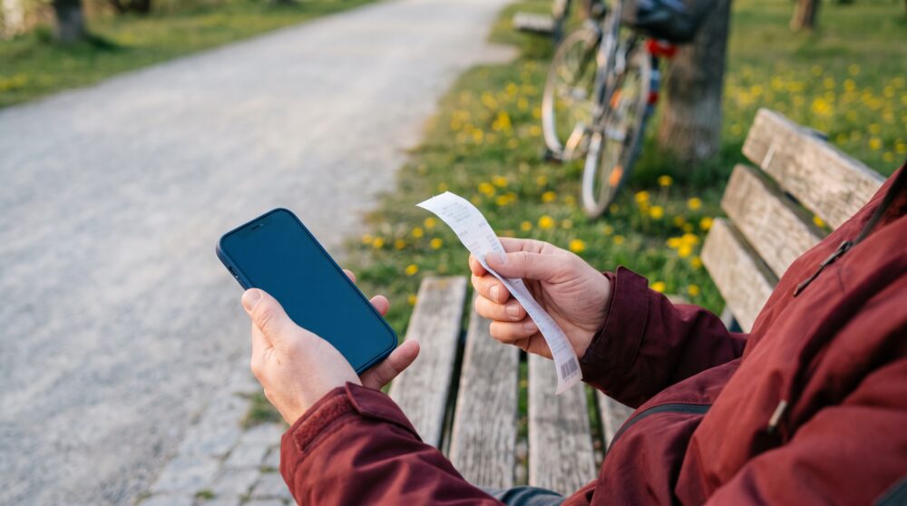 Person auf einer Parkbank mit Smartphone und gefaltetem Kassenbon in den Händen