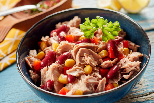 Thunfischstücke, rote Kidneybohnen, Mais und gewürfelte rote Paprika liegen in einer dunkelblauen Schüssel mit Petersilie.