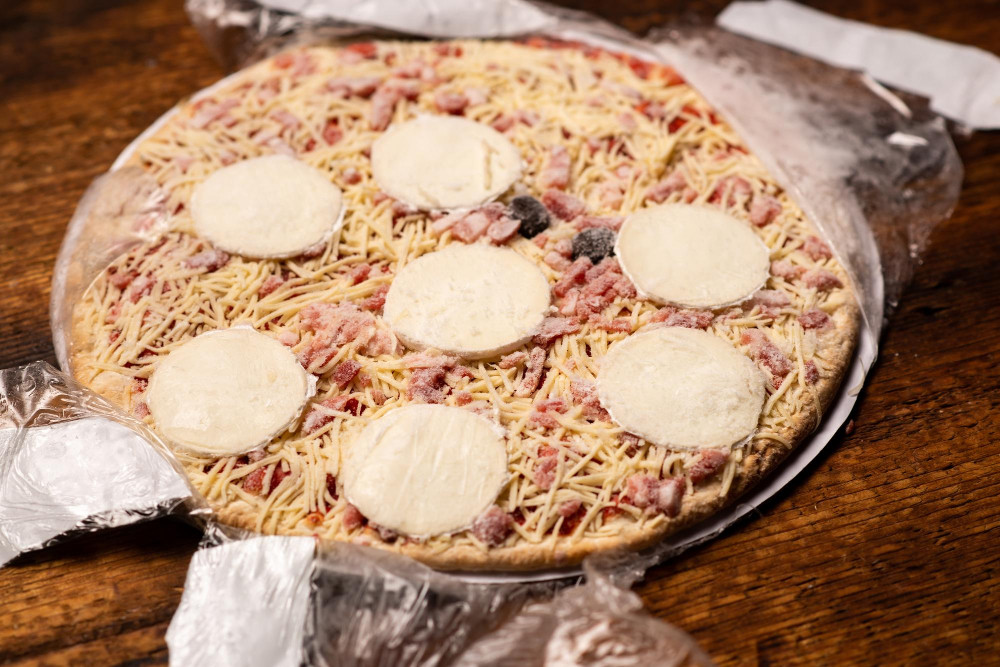Eine Nahaufnahme einer rohen Tiefkühlpizza auf einem Holztisch, die teilweise aus ihrer Plastikfolie ausgepackt ist. Der Belag besteht aus geriebenem Käse und mehreren runden Mozzarellascheiben.