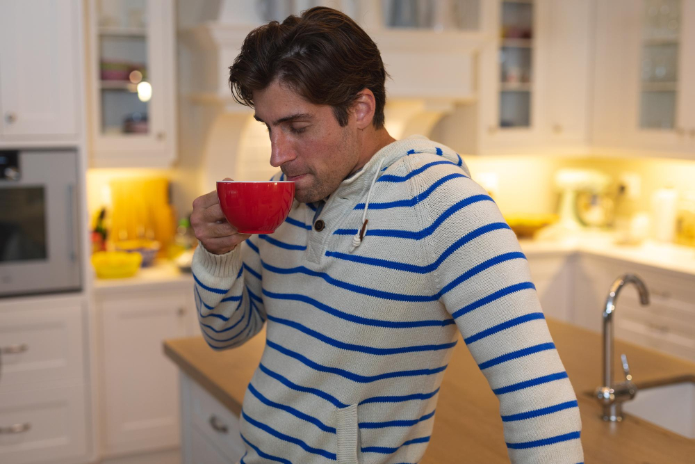 Mann in gemütlichem Pullover, der in der Küche aus einer roten Tasse trinkt – ein Moment der Ruhe, um Tipps gegen Motivationslosigkeit am Montag zu veranschaulichen.