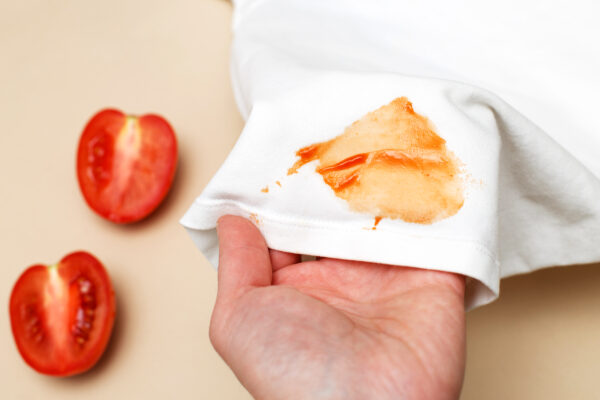 Eine Hand hält weißen Stoff mit orange-rotem Fleck; daneben liegen zwei aufgeschnittene Tomatenhälften.
