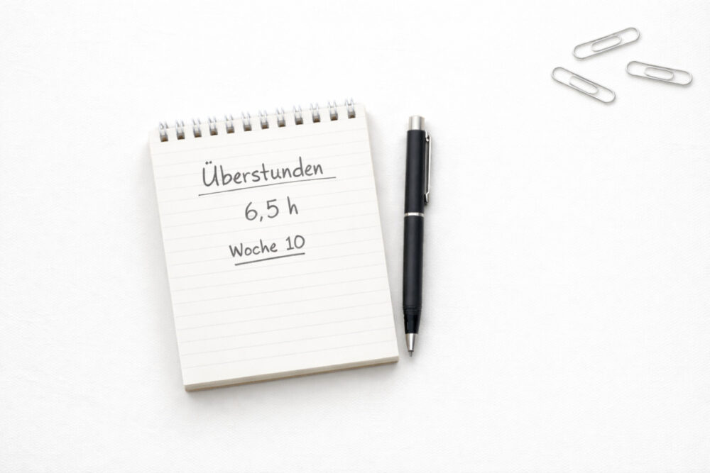 Notizblock mit Eintrag „Überstunden 6,5 h, Woche 10“, daneben ein schwarzer Stift und kleine Büroklammern auf hellem Schreibtisch
