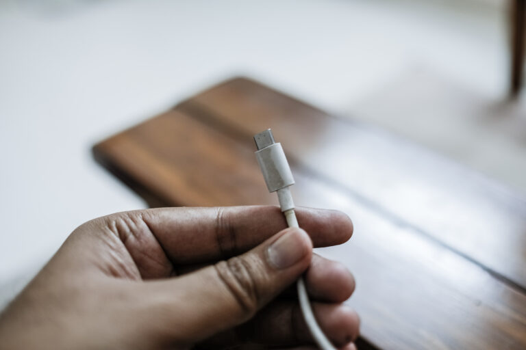 Eine Hand hält einen USB-C-Stecker vor einem unscharfen braunen Gegenstand auf einer hellen Fläche.