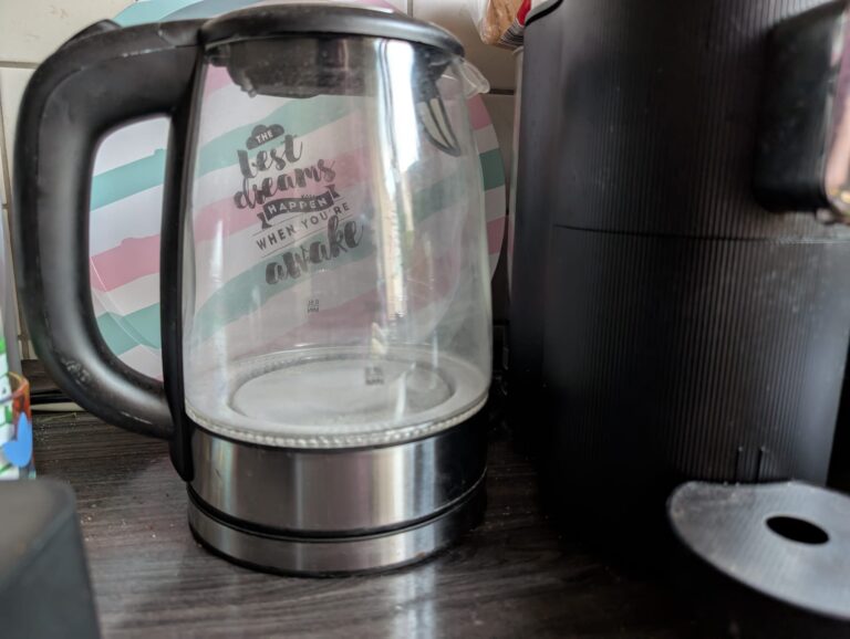 Verkalkter Glaswasserkocher vor bunt gestreiftem Tablett mit Spruch