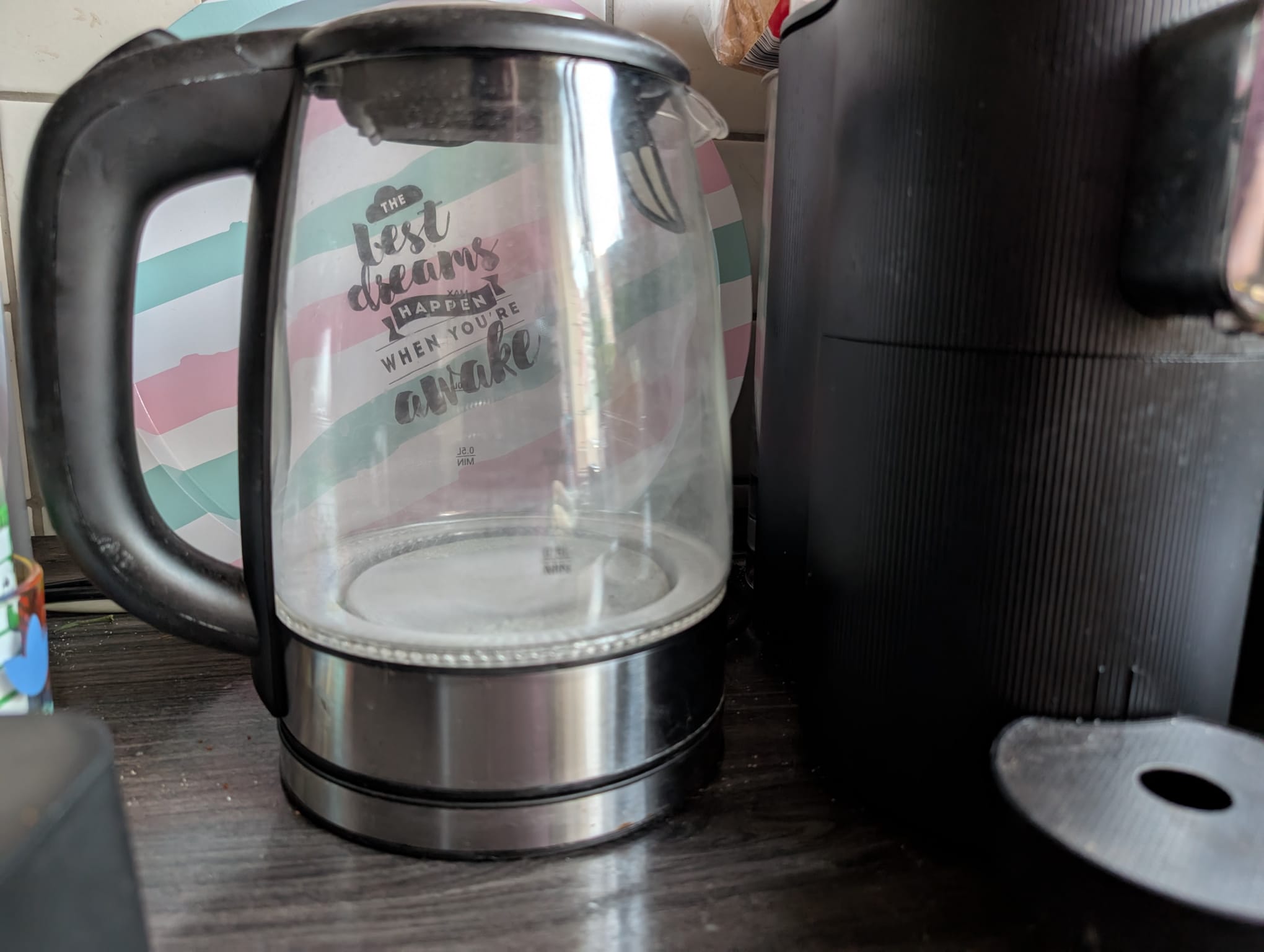 Verkalkter Glaswasserkocher vor bunt gestreiftem Tablett mit Spruch