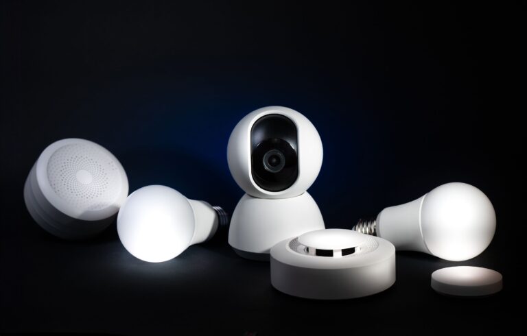 Mehrere weiße Smart-Home-Geräte stehen vor dunklem Hintergrund, darunter eine Kamera, runde Sensoren und zwei Lampen.