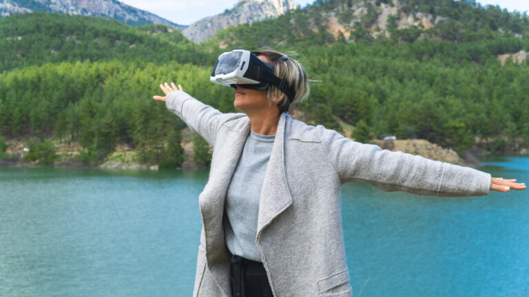 Eine Frau mit VR-Headset steht mit ausgebreiteten Armen vor einem türkisfarbenen See, Wäldern und Bergen.