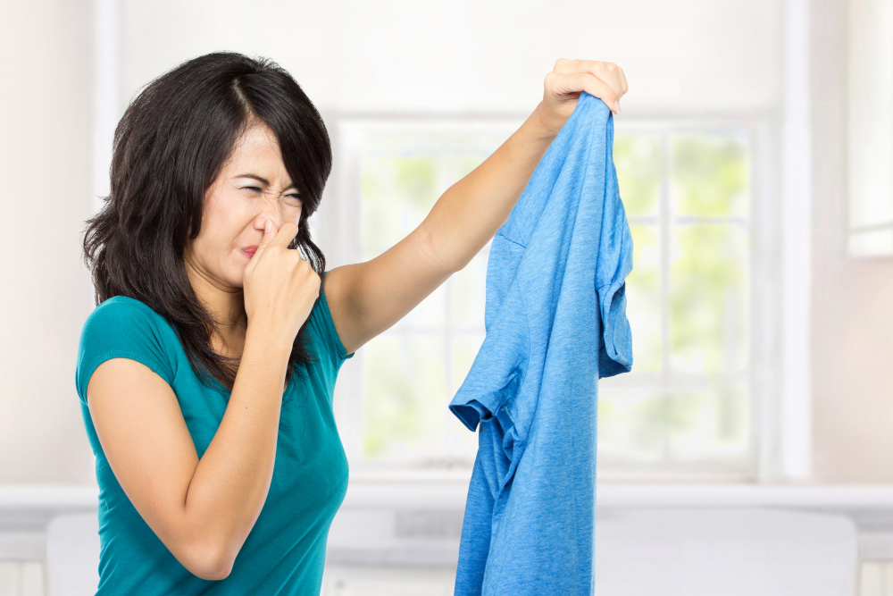 Frau hält sich die Nase zu und hält stinkendes blaues T-Shirt hoch