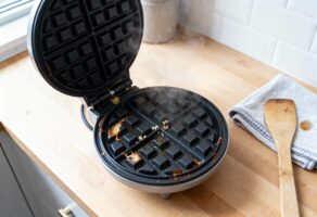 Rundes Waffeleisen mit leichten eingebrannten Teigresten, noch dampfend; daneben Mikrofasertuch und Holzspatel - Waffeleisen reinigen im warmen Zustand.