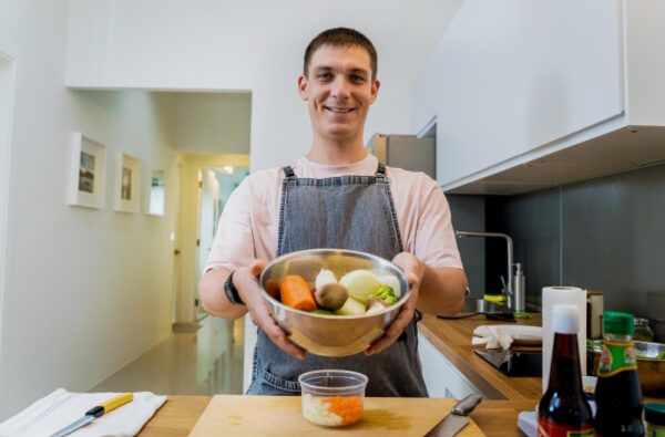 Ein Mann in einer modernen Küche hält eine Schüssel mit frischem Gemüse wie Karotten, Zwiebeln und Kartoffeln in der Hand und lächelt in die Kamera. Das Bild könnte symbolisieren, dass Clean Eating bedeutet, auf frische, unverarbeitete Zutaten zu setzen, die direkt in der Küche selbst zubereitet werden.