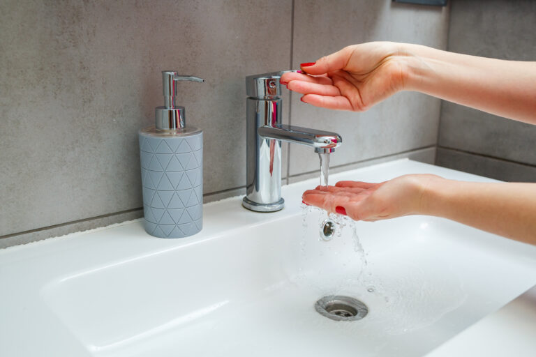 Zwei Hände halten sich unter einem laufenden verchromten Wasserhahn über einem weißen Waschbecken.