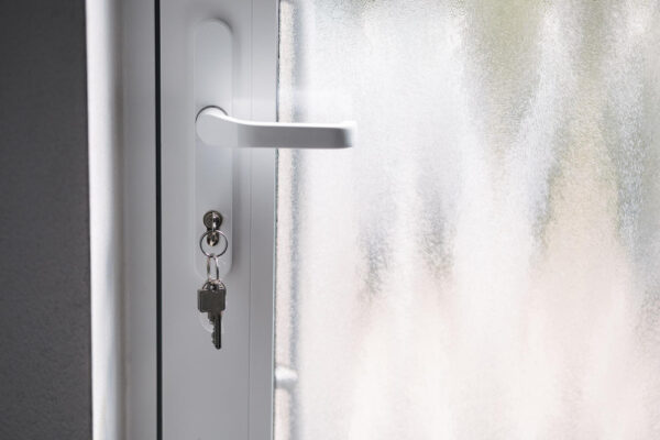 Detailansicht eines weißen Türgriffs mit einem Schlüsselbund, der an einem im Schloss steckenden Schlüssel hängt, vor einem Mattglaspaneel.