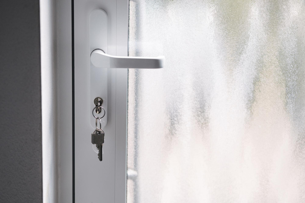 Detailansicht eines weißen Türgriffs mit einem Schlüsselbund, der an einem im Schloss steckenden Schlüssel hängt, vor einem Mattglaspaneel.