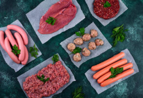 Welches Fleisch ist am ungesündesten - Auswahl aus Wurst, Hackfleisch, Steaks und Fleischbällchen auf Backpapier mit Kräutern, Draufsicht auf dunklem Untergrund.