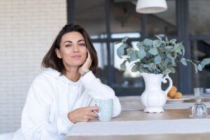 Frau in weißem Hoodie sitzt beim Wellness zu Hause entspannt mit Tasse am gedeckten Tisch