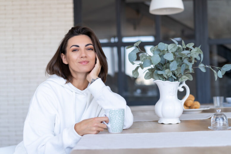 Frau in weißem Hoodie sitzt beim Wellness zu Hause entspannt mit Tasse am gedeckten Tisch