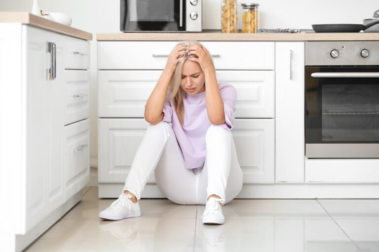 Frau sitzt erschöpft am Küchenboden und hält den Kopf