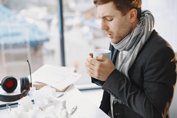 Mann sitzt frierend am Schreibtisch im Büro, trägt Schal und hält eine warme Tasse; Taschentücher und Kopfhörer liegen daneben.