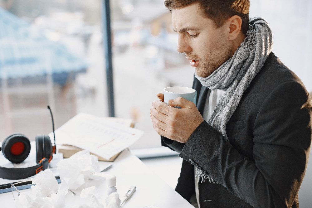 Mann sitzt frierend am Schreibtisch im Büro, trägt Schal und hält eine warme Tasse; Taschentücher und Kopfhörer liegen daneben.