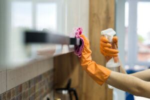 Eine Person mit orangefarbenen Gummihandschuhen reinigt einen weißen, glänzenden Küchenschrank. In der einen Hand hält sie eine Sprühflasche mit Reinigungsmittel, mit der anderen wischt sie mit einem rosa Mikrofasertuch über den schwarzen Griff des Schranks.