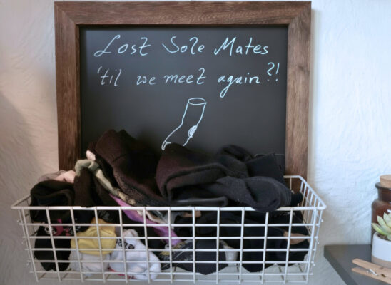Wohin verschwinden Socken: Drahtkorb mit einzelnen Socken vor Kreidetafel „Lost Sole Mates“