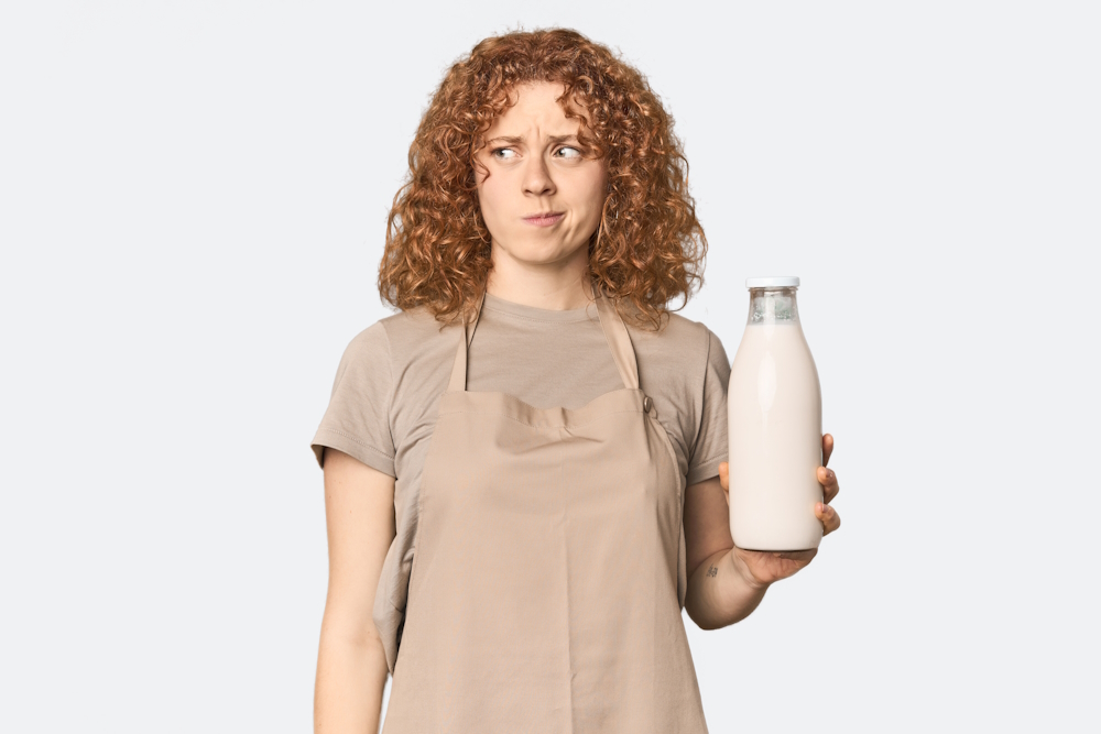 Junge kaukasische rothaarige Frau mit einer Milch in der Hand, die verwirrt ist, sich unsicher fühlt und zweifelt als Symbolbild für den Ratgeber: Schlechte Milch getrunken