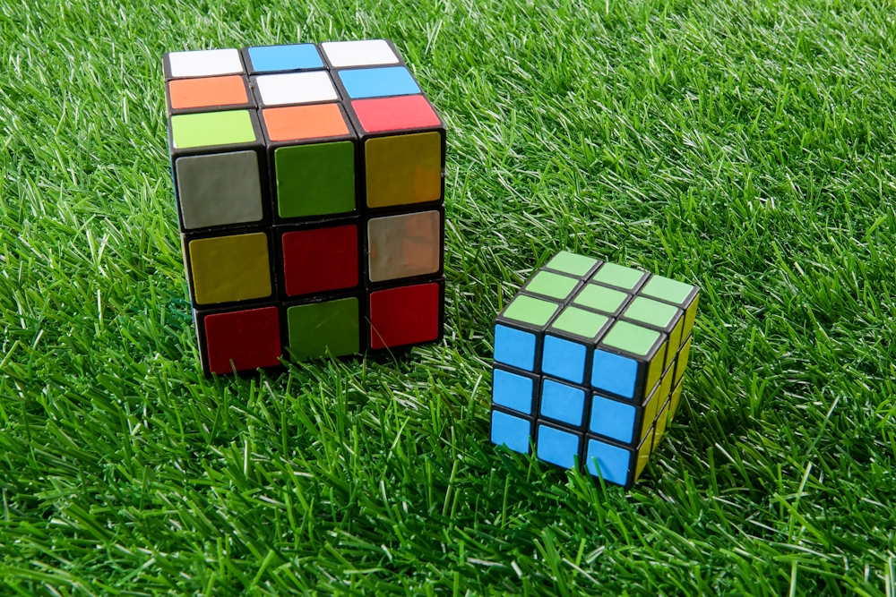 Zwei unterschiedlich große Rubik’s Cubes auf grünem Rasen, einer gelöst, einer ungeordnet.