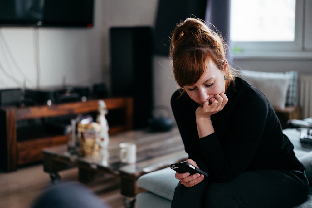 Frau sitzt nachdenklich mit Smartphone auf dem Sofa in einem Wohnzimmer.